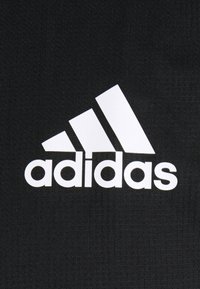 Logo Adidas blanc avec trois bandes diagonales au-dessus du nom de la marque sur un fond en tissu noir texturé.
