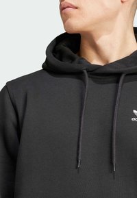 Svart hoodie av mjukt tyg, med huva med dragsko, ribbade ärmslut och en liten vit logga på bröstet.