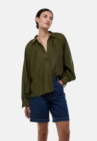 Chemise boutonnière olive, ample, avec col, épaules froncées et manches longues ; associée à un short en denim noir.