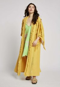 Geel franjes lange kimono gedragen over een groene jurk met roze bloemenpatroon. Model staat op een effen achtergrond en toont de vloeiende uitstraling van het kledingstuk.