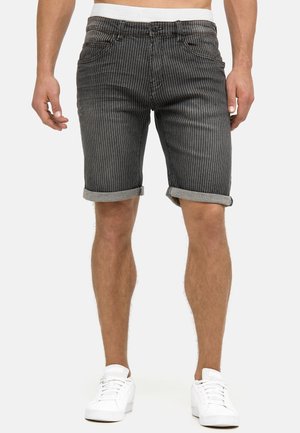 Mann, der schwarze, schwarz gestreifte, hochgekrempelte Shorts und weiße Turnschuhe trägt, vor weißem Hintergrund steht.