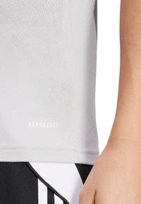 Lichtgrijs sportshirt met textuurstof; voorzien van het "AEROREADY"-logo aan de zoom. Zwarte shorts met witte accenten zijn ook zichtbaar.