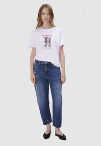 T-shirt blanc avec imprimé graphique et jean bleu usé, associé à des ballerines noires. Le mannequin pose devant un fond uni.