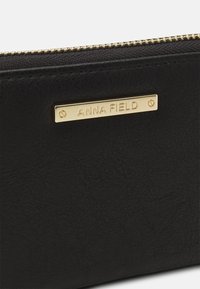 Schwarzes Kunstlederportemonnaie mit strukturiertem Oberflächenmaterial, ausgestattet mit einer goldfarbenen Metall-Logo-Plakette mit der Aufschrift "ANNA FIELD" und einem Reißverschluss.