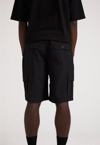 Schwarze Cargo-Shorts aus leichtem, strukturiertem Stoff. Mit Seitentaschen und Knopfverschluss. Tragen Sie dazu ein übergroßes schwarzes T-Shirt.