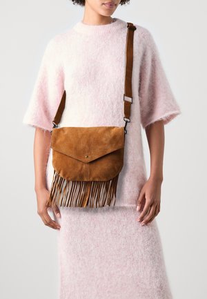 Femme portant une robe en maille pelucheuse rose et un sac bandoulière en suède marron avec des détails à franges et une sangle réglable.