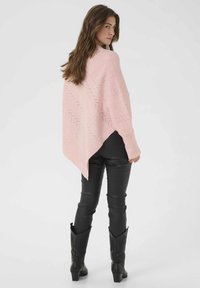 Rožinė nėriniuota pončo su zigzago raštu, pasižyminti asimetriniu kraštu. Modelis dėvi juodas aptemptas kelnes ir šikšnosparnių batus.