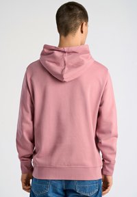 Sudadera rosa con capucha ajustable, mangas largas y puños de canalé. Tela suave con una textura lisa. Combinada con jeans de mezclilla azul.