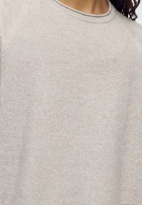 Gros plan d'une personne portant un pull en maille beige avec un col rond et des manches raglan.