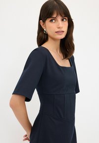 Vestito blu navy con maniche corte e scollatura quadrata. Presenta pannelli strutturati e una texture liscia, progettato per una silhouette aderente.