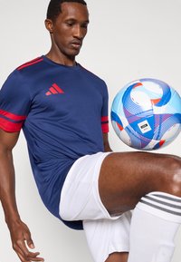 Blaues Adidas-Sportshirt mit roten Akzenten, kombiniert mit weißen Shorts und gestreiften Socken, während ein bunter Fußball auf dem Oberschenkel balanciert wird.