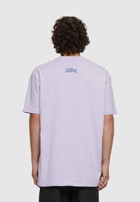 Světle levandulové bavlněné tričko s krátkým rukávem a volným střihem; obsahuje malé modré grafické logo na horní části zad.