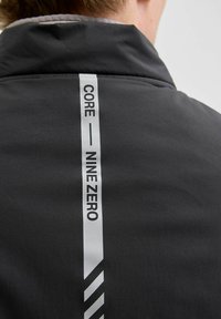 Czarna kurtka o teksturowej powierzchni, z pionowym białym paskiem z logo oznaczonym "CORE" i "NINE ZERO" z akcentem w zygzak.