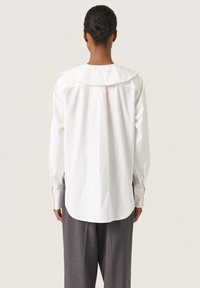 Camicia bianca a maniche lunghe con colletto arricciato, piega sul retro e tessuto liscio. Indossata con pantaloni grigi a righe.