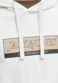 Lichtgrijze hoodie met een voorzak, voorzien van een groot logo in beige en zwart. De tekst luidt "JACK & JONES" en "SINETEEN NINETY."