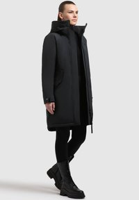 Parka aislante negra con un cuello alto, cremallera frontal, bolsillos laterales y puños ajustables, combinada con botas negras de cordones.
