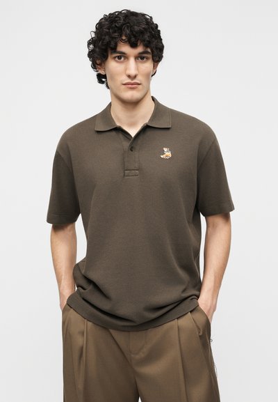 Άνδρας με σγουρά σκούρα μαλλιά, φοράει καφέ κοντομάνικο πόλο με ένα μικρό έμβλημα αλεπούς και καφέ πλισέ παντελόνια, με τα χέρια στις τσέπες.