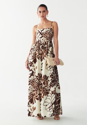 Frau trägt ein ärmelloses, bodenlanges Kleid mit großem braunem Blumenmuster auf cremefarbenem Hintergrund, hält eine beige Clutch und trägt goldene Ohrringe.