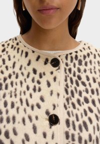 Leopardí fleecová bunda s kulatým výstřihem, béžovým lemem a černými knoflíky. Měkká textura a výrazné skvrny v tmavě hnědé na krémovém pozadí.