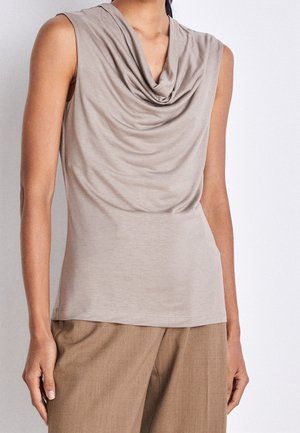 Beżowa bluzka bez rękawów z drapowanym dekoltem noszona z jasnobrązowymi spodniami na stojącej osobie na jednolitym tle.