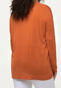 Luźna, długosleeve bluzka w kolorze pomarańczowym, wykonana z miękkiego materiału. Posiada szeroki dekolt i ściągacz na dole, zestawiona z białymi spodniami.