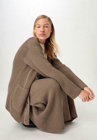 Gestreifter brauner Cardigan mit langen Ärmeln, seitlichen Taschen und lockerer Passform, getragen über einem beigen Oberteil, hervorgehoben vor einem neutralen Hintergrund.