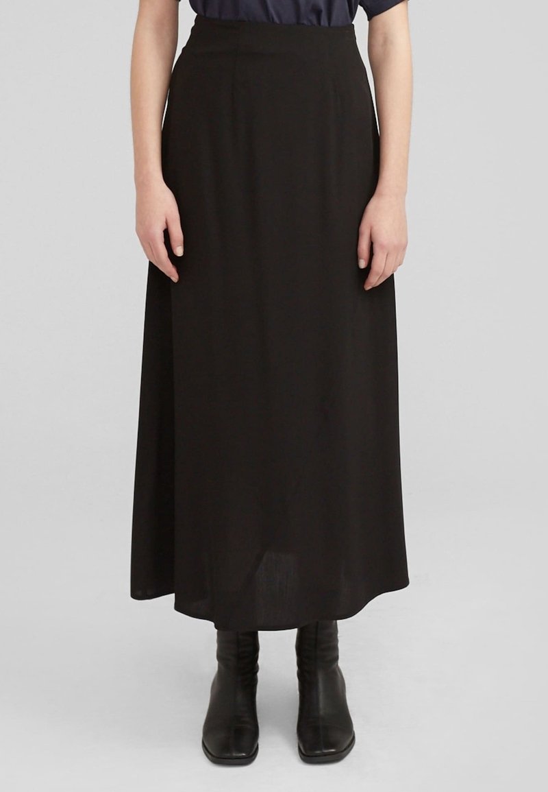 Femme portant une jupe noire longueur cheville, des bottines noires à bout carré et un haut bleu foncé, debout devant un fond gris uni.