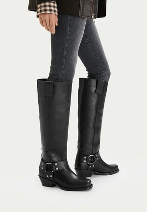 Nogi w ciemnoszarych obcisłych dżinsach, czarne skórzane botki do kolan z srebrnymi klamrami oraz czarna kurtka z widocznymi mankietami koszuli w kratę.