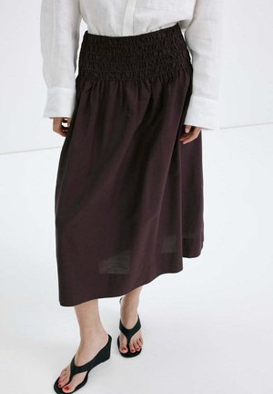 Mujer con blusa blanca de manga larga, falda midi negra de cintura alta y sandalias de cuña negras con esmalte de uñas rojo en los dedos de los pies.