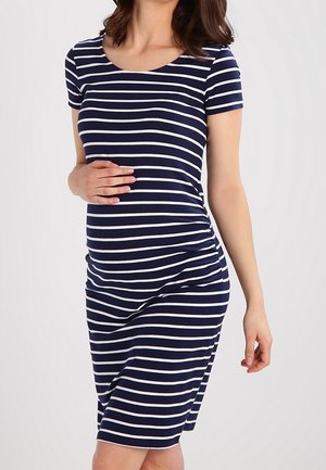 Zwangere vrouw die een marineblauw-wit gestreepte jurk met korte mouwen draagt en zachtjes met één hand haar buik vasthoudt.