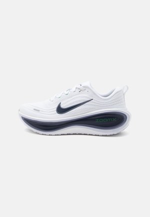Baskets blanches de sport avec swoosh Nike noir et semelle épaisse incurvée noire et blanche labellisée ZoomX, montrées de profil sur fond uni.