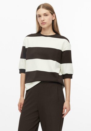 Femme aux cheveux lisses châtain clair portant un haut à manches courtes rayé large noir et blanc et un pantalon foncé sur fond blanc.