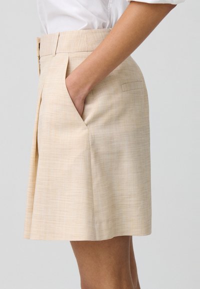 Gonna beige al ginocchio realizzata in tessuto texturizzato. Presenta tasche laterali e una vita classica con pieghe per un tocco di dettaglio in più.