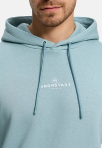 Ljusblå huvtröja i mjuk bomullsblandning, med framfickor i kängurustil, dragsnören och en vit logotyp på bröstet.