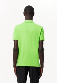 Osoba s tmavou pletí, oblečená v polo tričku s krátkým rukávem limetkové barvy a černých kalhotách, zobrazená zezadu na bílém pozadí.