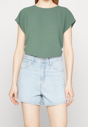 Personne portant un haut vert ample à manches courtes et un short en jean bleu clair taille haute, les mains détendues le long du corps.