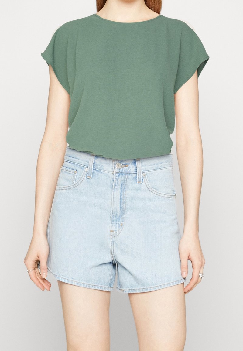 Personne portant un haut vert ample à manches courtes et un short en jean bleu clair taille haute, les mains détendues le long du corps.