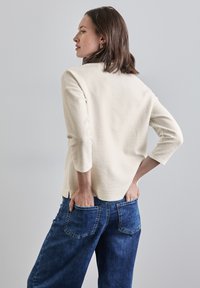 Cremfarbener Pullover mit 3/4-Ärmeln, kombiniert mit lockeren blauen Jeans, die über Taschen auf der Rückseite und einen lässigen Schnitt verfügen.