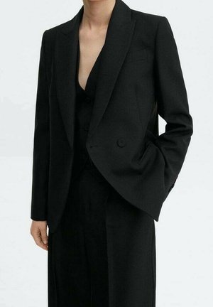 Henkilö, joka pukeutuu räätälöityyn mustaan kaksiriviseen bleiseriin ja vastaaviin mustiin housuihin, toinen käsi taskussa, vaaleaa taustaa vasten.