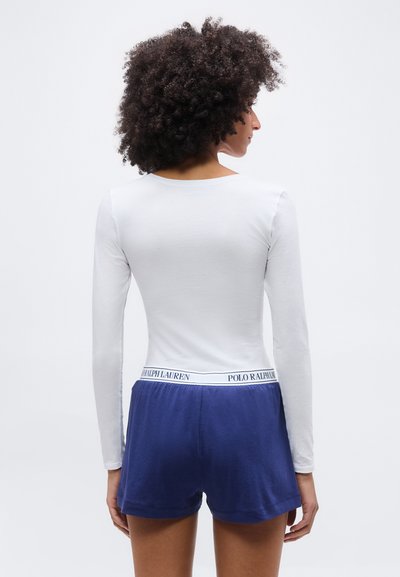 Γυναίκα με σγουρά μαλλιά, φορώντας λευκή μπλούζα μακρυμάνικη και σκούρα μπλε σορτς με ζώνη Polo Ralph Lauren, στέκεται με την πλάτη προς τα έξω.