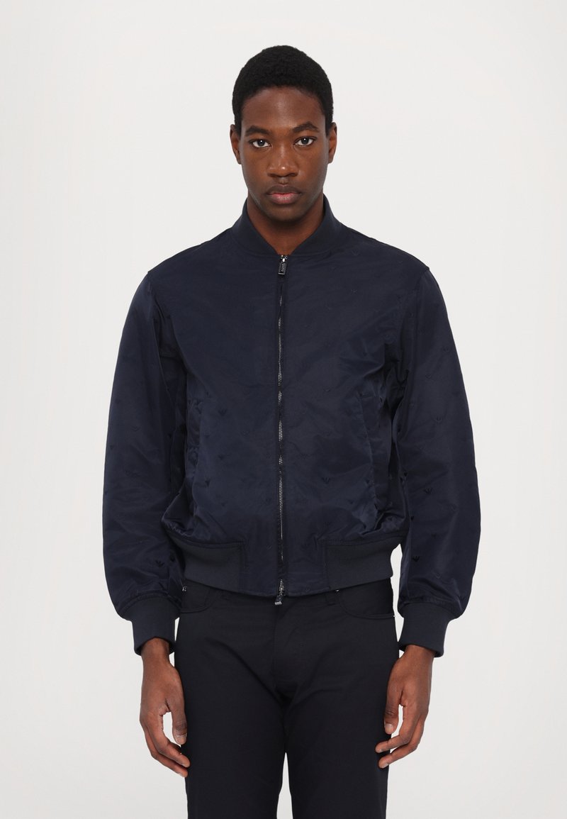 Navy bomber jacket with a zip front, ribbed cuffs, and waistband. Smooth, lightweight fabric features subtle embossed pattern.