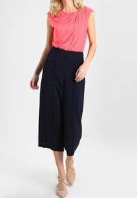 Top s korálovými závěsy a krátkými rukávy v kombinaci s námořnickými kalhotami culottes s vysokým pasem; modelka má na sobě nahé sandály na tenké podrážce, čímž vytváří čistý a strukturovaný vzhled.