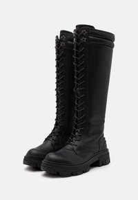 Černé kožené kozačky sahající po kolena s kulatou špičkou, výrazným šněrováním vpředu, hvězdovitými kovovými ozdobami a masivními gumovými podrážkami.