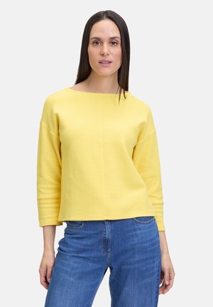 Pullover giallo con trama, scollatura a barca e maniche a tre quarti, abbinato a jeans blu. Tessuto morbido, vestibilità rilassata.