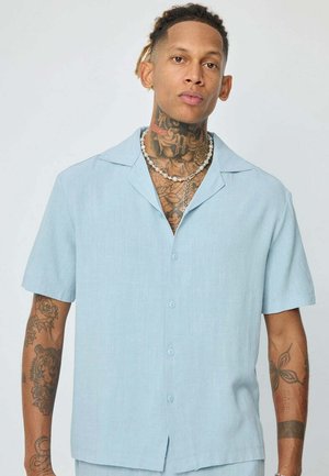 Homme avec des tatouages au cou et au bras, portant une chemise bleu clair à manches courtes boutonnée et un collier de perles sur un fond uni.
