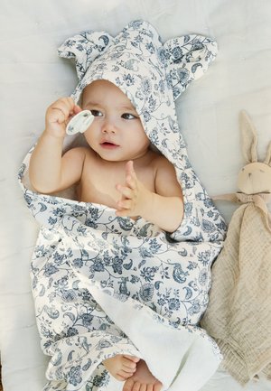 Vauva, joka on kääritty kukikkaaseen hupulliseen pyyhkeeseen ja pitää tuttia, makaa tilkkupeitolla beigen pehmojäniksen vieressä.