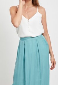 Frau in einem weißen ärmellosen Oberteil mit Knöpfen, kombiniert mit einem hochtaillierten, plissierten, hellblauen Rock vor einem schlichten Hintergrund.