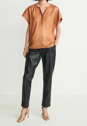 Femme portant un chemisier court en satin cuivre, un pantalon en cuir noir, des sandales à talons hauts couleur nude, se tenant devant un fond blanc uni.