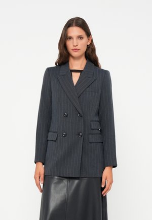 Dobbeltradet mørkegrå blazer med hvite pinstriper, sklippeslag, to frontlommer og rundede skulderlinjer, båret over et svart skjørt.
