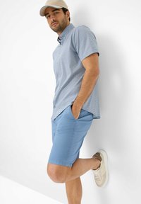 Camicia azzurro chiaro a maniche corte con un motivo testurizzato, abbinata a pantaloni corti azzurro chiaro e sneaker beige. Uomo appoggiato a un muro bianco.